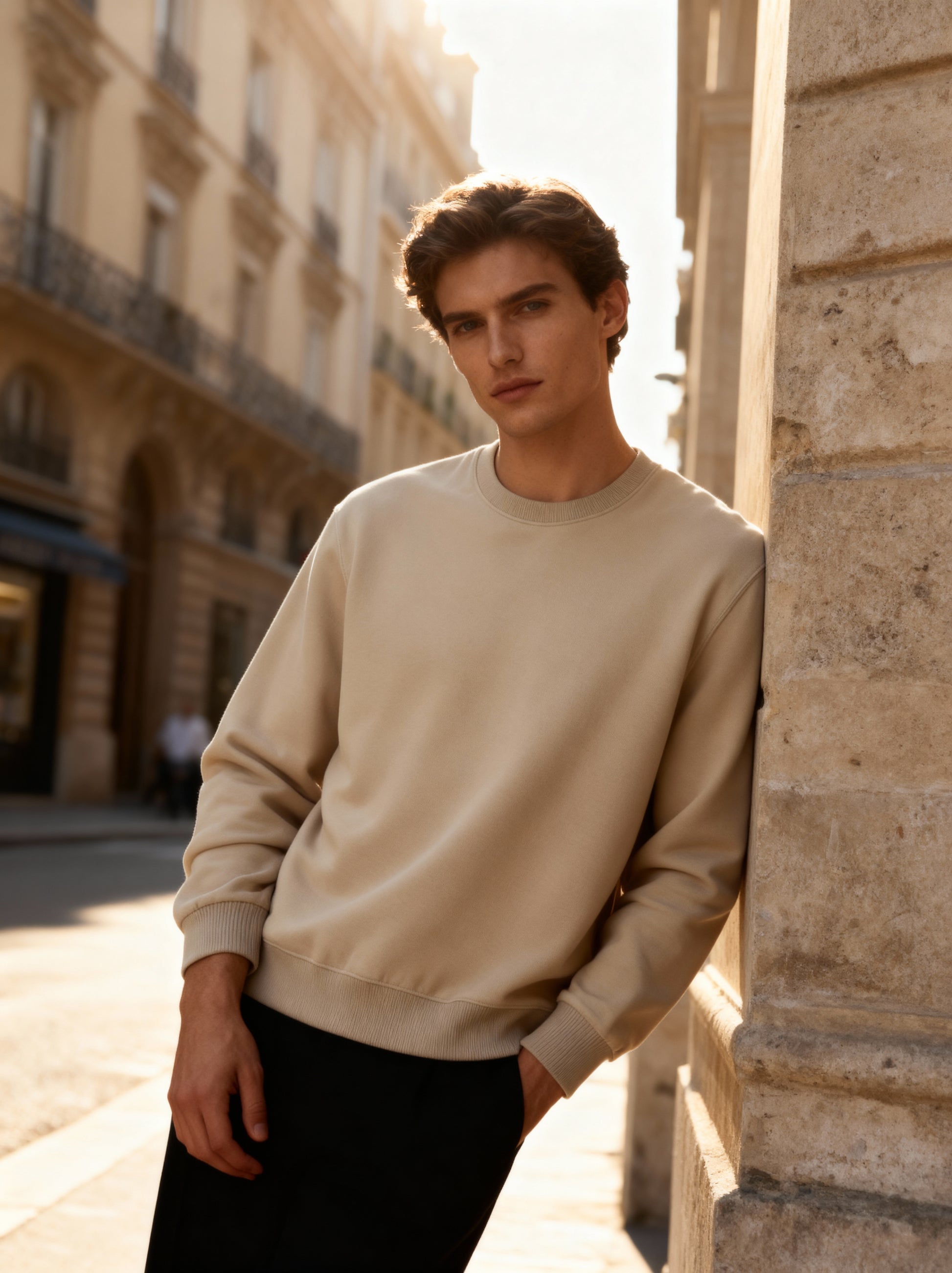 Man wearing a beige sweater leaning against a stone wall in an urban setting