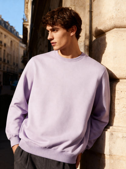 Man wearing a light purple sweatshirt standing against a stone wall in an urban setting.