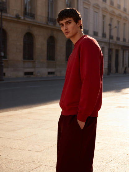 Man wearing a red sweatshirt and dark pants standing on a street with buildings in the background aaploo luxury